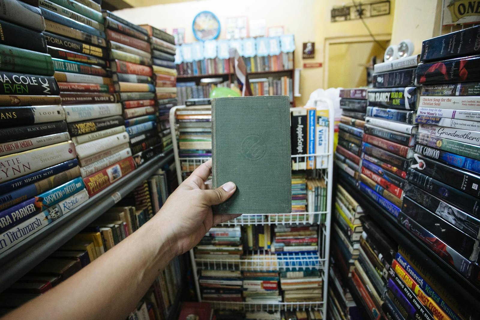 A hand holds a hardcover book amidst stacks of books in a vibrant, cozy bookstore.
